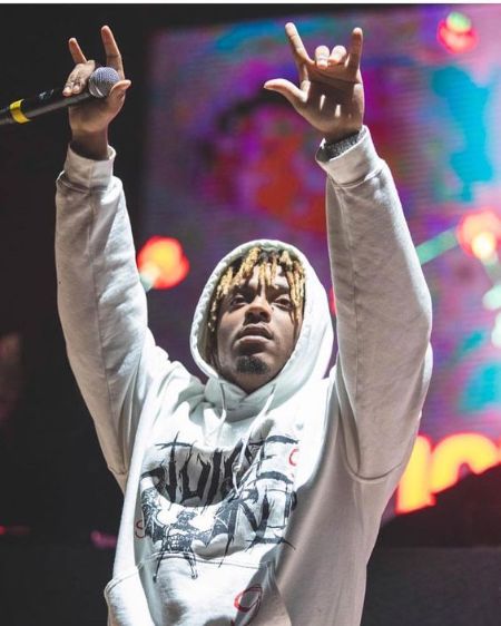 juice Wrld on stage wearing a white hoodie with his hands up in the air holding a mic. 